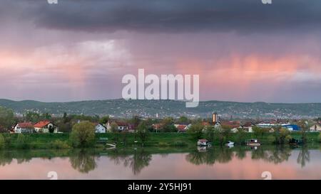 Strong downpour over hill behind settlement, sunlit clouds during sunset Stock Photo
