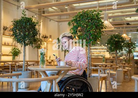 Thoughtful dreamy disabled man in wheelchair drinking coffee at cafe table, using mobile phone Stock Photo