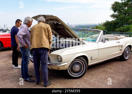 An outdoor exhibition of vintage cars. Three gentlemen checking out the engine of a vintage convertible Ford Mustang car. Stock Photo