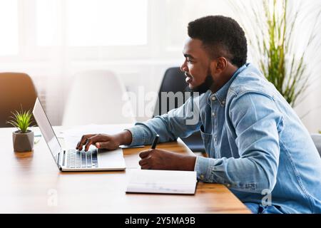 African worker taking notes while using laptop in office Stock Photo