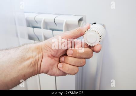 Male hand adjusting the thermostat knob on a white heater radiator, illustrating the control of indoor heating and temperature management in a home or Stock Photo