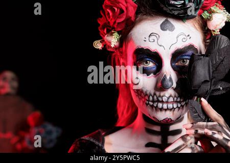 Headshot portrait of woman holding black rose set against dark background celebrating Dia de los Muertos holiday. Close up of lady with flower during Halloween horror event Stock Photo