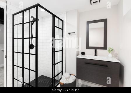 A bathroom with a floating wood cabinet, wooden mirror over a white sink, and a shower with white subway and black hexagon tiles. Stock Photo