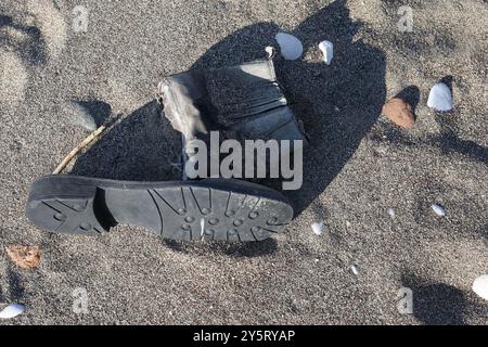 A single, worn-out black boot lies abandoned on a sandy beach. Small rocks and seashells are scattered around, under the strong sunlight of the coasta Stock Photo