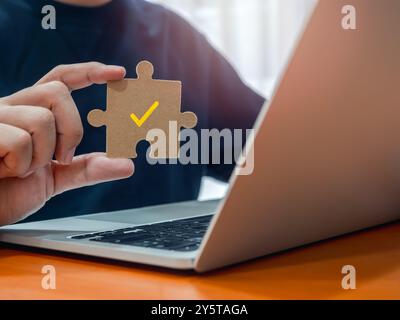 Yellow checkmark icon on jigsaw puzzle piece holding by businessman's hand while working with laptop computer at office. Business goal and success, so Stock Photo