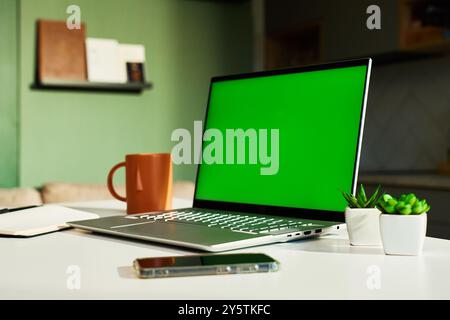 Modern home workspace featuring laptop with green screen, smartphone, coffee mug and small potted plants. Home office for freelancer with kitchen in b Stock Photo
