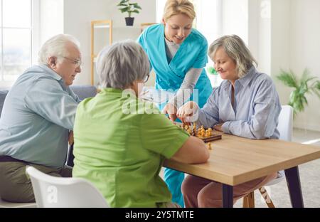 Young friendly female nurse or social worker helping senior people in playing chess in nursing home Stock Photo