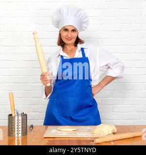 beautiful chef woman in uniform and hat with rolling pin in kitchen on brick wall background Stock Photo
