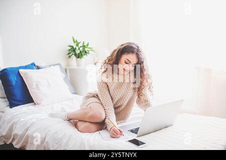 University student attending morning online class on laptop, sitting on bed in dorm room. Stock Photo