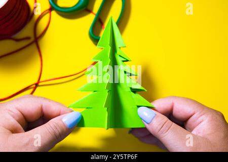 Female hands holding origami Christmas tree with a yellow background and on it are placed scissors and a Christmas decorative thread Stock Photo