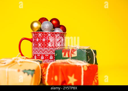 Christmas concept with multi-colored Christmas balls, with decorated red mug and gifts around on a yellow background Stock Photo
