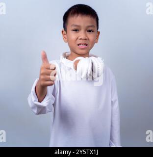 Asian boy wearing white with white headphones on his neck is giving thumbs up gesture showing excellent, Boy standing on white background, Lifestyle o Stock Photo