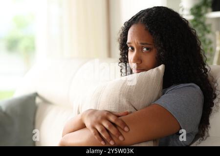 Sad black woman looks at you embracing pillow sitting on a couch at home Stock Photo