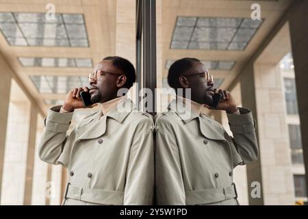 African American man standing next to large window, holding smartphone to ear while posing in trench coat, and looking at his reflection Stock Photo