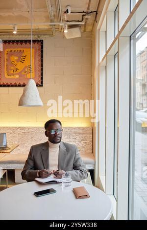 Man wearing sunglasses and beige turtleneck sweater writing notes while sitting at a table by large window in cafe. Modern interior with artistic decor on wall present Stock Photo