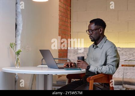 Man wearing light gray shirt sitting at white desk working on laptop and holding reusable cup of coffee with empty brick wall in background Stock Photo