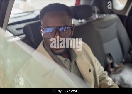 African American man sitting in car wearing sunglasses, looking thoughtfully out window, dressed in stylish beige coat and shirt, interior background of vehicle highlighted Stock Photo