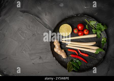 Ingredients for making Tom Yum Kung. Lemongrass, chili pepper, ginger, galangal, lime, cilantro and cherry tomatoes on a black plate against a dark ba Stock Photo