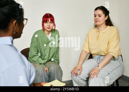 Three individuals engaging in therapy session, including therapist and two women on chairs. One woman has red hair while the other has brown hair, both wearing casual clothes Stock Photo