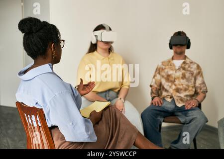 Three people engaging virtual reality experience in education setting with headsets while sitting in chairs, each exploring various applications and environments in immersive learning Stock Photo