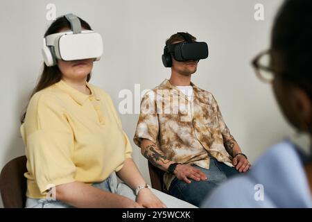 Individuals immersed in virtual reality experiences, wearing VR headsets in minimalist room with plain wall background. Engaging with advanced technology, creating unique virtual experiences Stock Photo