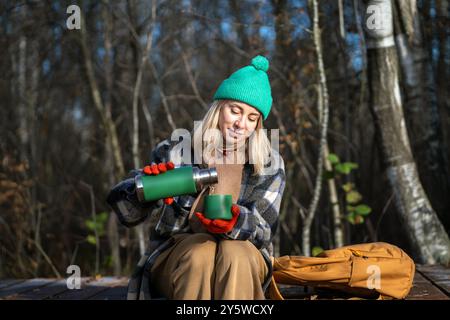 Woman hiker having break drinking tea sitting on wooden path in autumn park in Scandinavia. Stock Photo