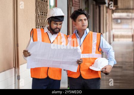 Two engineers are seen working together at a construction site, engaged in a detailed discussion about the project floor plan and blueprint. Both are Stock Photo