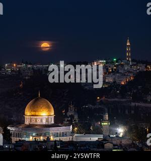 A full, orange moon rises low on the horizon over Mount Scopus, the Dome of the Rock and the Old City of Jerusalem, Israel. Stock Photo