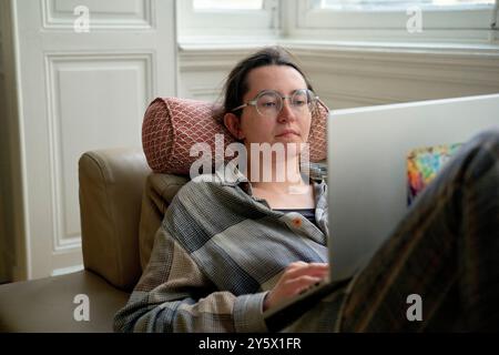 A focused adult wearing glasses and a checkered shirt is working on a laptop while reclining comfortably on a beige armchair. Stock Photo