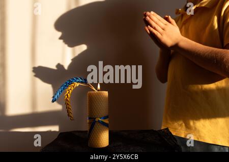 burning candle, two ears of wheat, painted yellow-blue against of shadow of child with his hands folded in prayer Stock Photo