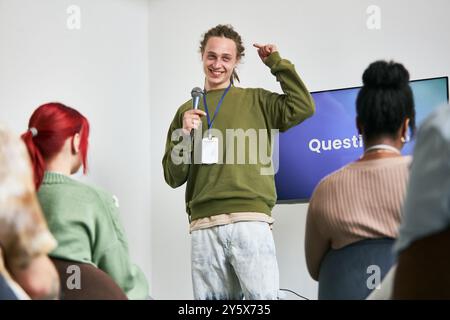 Young man standing with microphone smiling and interacting with diverse audience in indoor setting during presentation session Stock Photo