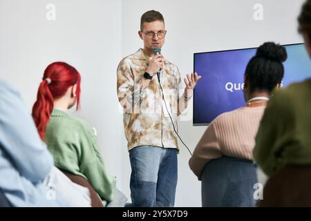 Presenter standing in front of group of diverse people, explaining ideas with microphone and pointing to screen. Audience sitting attentively, focused on presenter Stock Photo