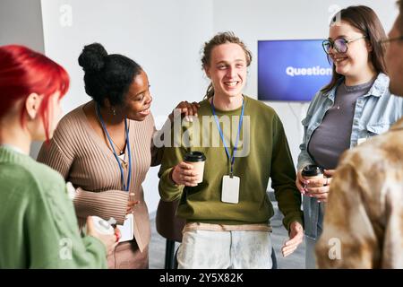 Group of young adults from diverse backgrounds discussing ideas at informal meeting while holding coffee cups and wearing casual clothing. Smiling and having conversation Stock Photo