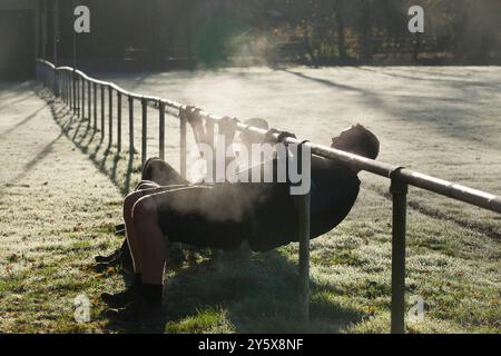 A person exercises on an outdoor bar on a misty morning, with the sun shining through the trees and illuminating the dew-covered grass. Stock Photo