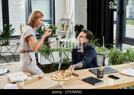 Two young colleagues enjoy lunch together, sharing pizza and smiles in a vibrant workspace. Stock Photo