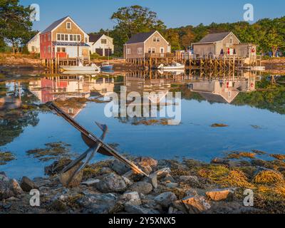 Coastal houses are reflected in calm water, with moored boats and an old anchor in the foreground. The scene is set against a backdrop of trees, creat Stock Photo