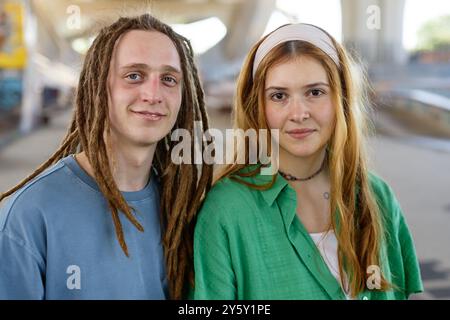 Portrait of young couple with dreadlocks and casual outfits standing under overpass smiling together under shaded urban environment Stock Photo