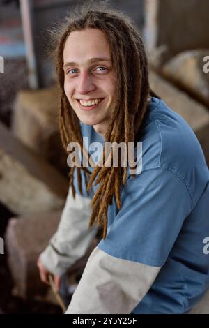 Young man with dreadlocks smiling while sitting on rocky surface and wearing a blue shirt and light jacket, creating an informal and relaxed environment Stock Photo