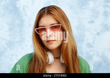 Portrait of young Hispanic woman with long hair wearing pink glasses and white headphones, standing against blue textured background Stock Photo