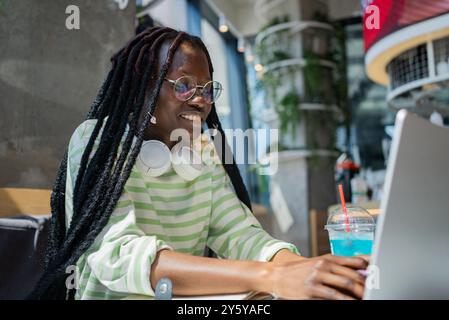African American woman with headphones around her neck uses a laptop in a modern cafe, enjoying a refreshing blue drink.  Stock Photo