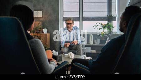 Mature psychotherapist talks to young family, provides couples therapy. Back view of multiethnic couple sitting on chairs, listening psychologist on counseling session. Psychology and sexology concept Stock Photo