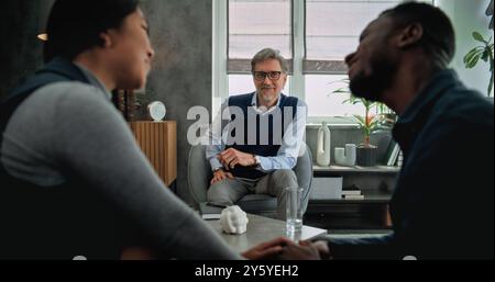 Happy mature psychotherapist talks to young family, provides counseling session. Back view of loving multiethnic couple sitting on chairs holding hands and laughing after quarrel on couples therapy. Stock Photo