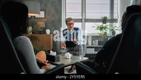 Mature psychotherapist talks to young family, provides couples therapy. Back view of multiethnic couple sitting on chairs, listening psychologist on counseling session. Psychology and sexology concept Stock Photo