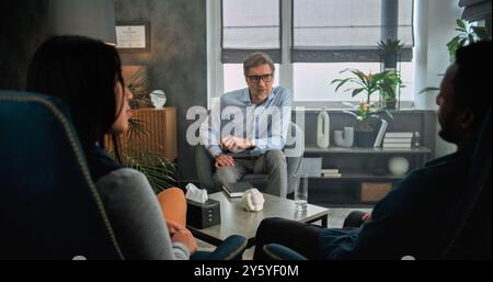 Mature psychotherapist talks to young family, provides couples therapy. Back view of multiethnic couple sitting on chairs, listening psychologist on counseling session. Psychology and sexology concept Stock Photo
