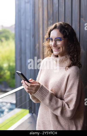 Smiling woman using smartphone, standing on balcony, enjoying leisure time Stock Photo