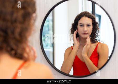 Looking in mirror, woman in red dress touching face, practicing self-care Stock Photo