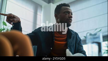Angry African American man loses his temper, starts fight and shouts during couples therapy. Multiethnic young couple having conflict on counseling session with family psychologist. Divorce process. Stock Photo