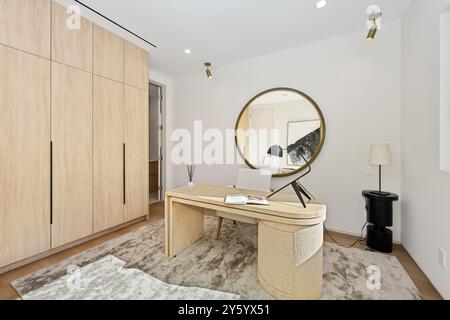 Modern home office with a minimalist design, featuring a wooden desk, round mirror, and ample storage space. Stock Photo