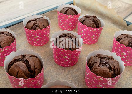 Brown chocolate muffins wrapped in white paper baked in the oven, lying on baking paper. Stock Photo