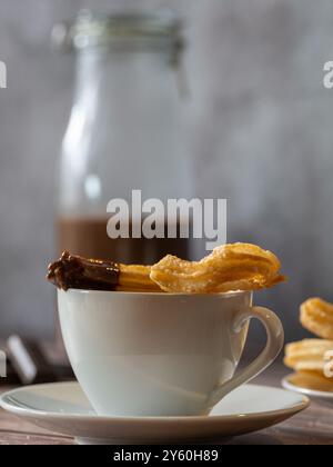 Closeup of a chocolate-covered churro over a white cup of hot chocolate. Glass bottle with chocolate in the background Stock Photo